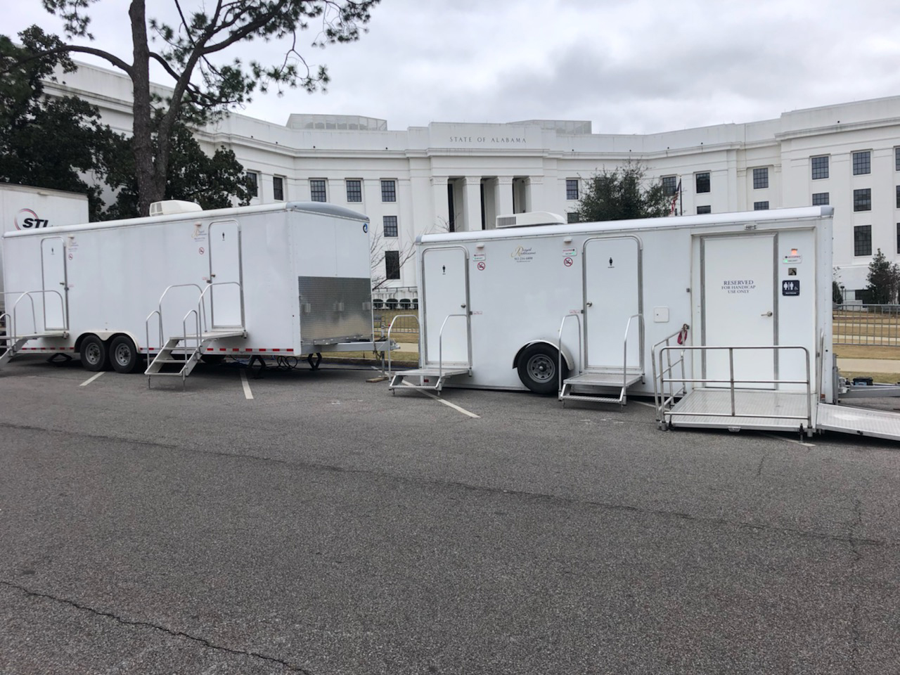 Two large white luxury portable restroom trailers parked in front of the State of Alabama building