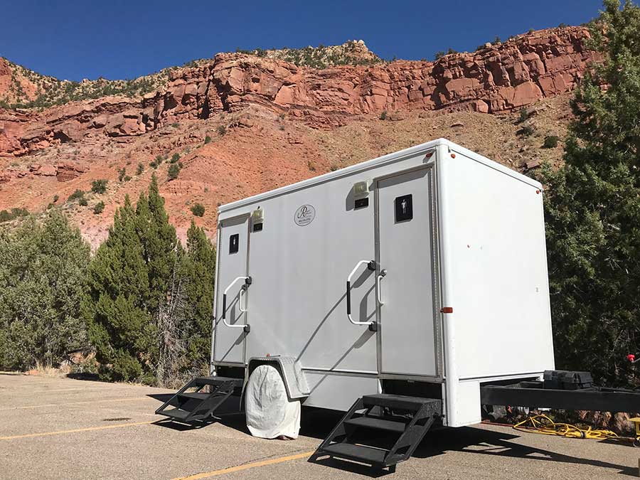 White portable restroom trailer with two doors and black stairs, parked in a lot with red rocky hills and green trees in the background under a clear blue sky