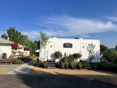 White portable restroom trailer with plants in black pots around the base, set outdoors on a sunny day with a clear blue sky.