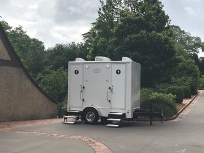 White Royal Restrooms portable restroom trailer with two doors labeled for men and women, placed on a paved area with trees and shrubs in the background on a cloudy day