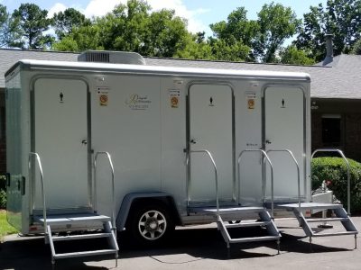 Silver portable restroom trailer with separate entry doors for men and women, each having steps and railings, parked on pavement with greenery and a building in the background.