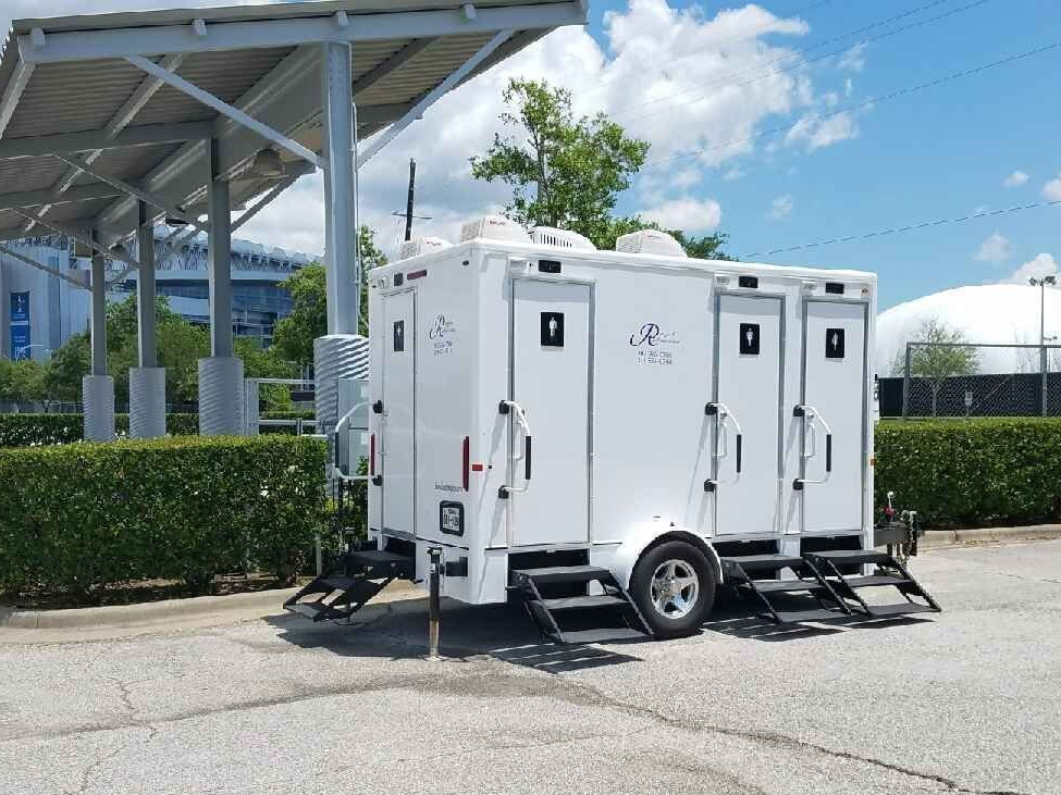White portable restroom trailer with four doors and three sets of steps, parked outdoors near hedges and metal structure.