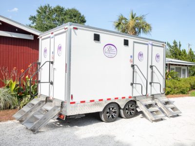 Portable white restroom trailer with dual separate entrances and small metal staircases on a sunny day.