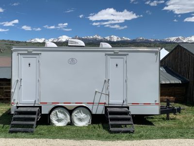 White portable restroom trailer with separate male and female entrances, black steps, and snow-capped mountains in the background.