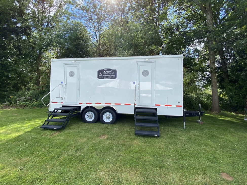 White portable restroom trailer with two doors and black steps on a grassy area surrounded by trees