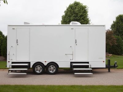 White mobile trailer with two doors, steps, and dual wheels parked on a paved surface with grass and trees in the background.