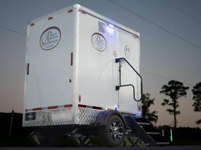 White portable restroom trailer with diamond plate lower panels, stairs leading to the door, and logos of Royal Restrooms on the side, set against a clear evening sky with silhouetted trees in the background.